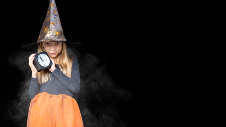 A happy smiling little girl in a carnival witch costume holds in her hands a black alarm clock on a...