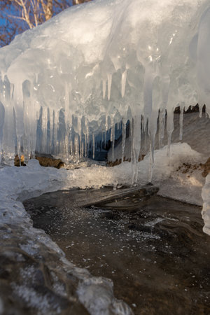 Rows of stalactite icicles hang from the stone, close-up, vertical frame. Icicles on the lake beach on the background of blue water. Fragments of ice on the surface of a frozen lake.の写真素材