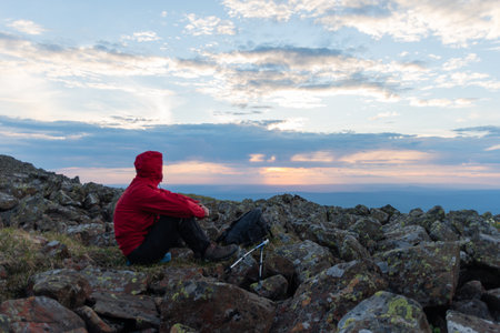 A man in a red jacket with a backpack and trekking poles enjoying the sunset at the top of the...