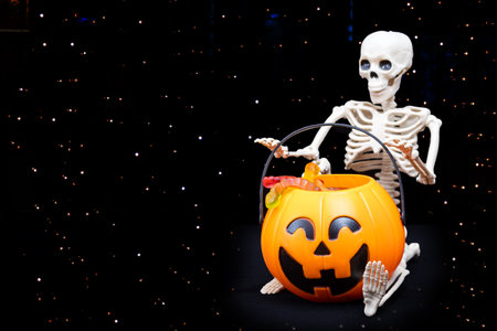 A sitting skeleton takes marmalade candies out of a bucket in the shape of a pumpkin, black background, copy space. Trick or treat for Halloweenの写真素材