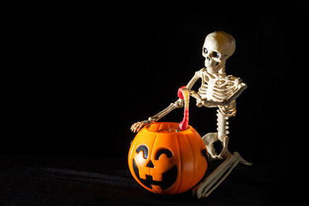 A seated skeleton pulls out of a bucket in the shape of a pumpkin candy on a black background, copy...