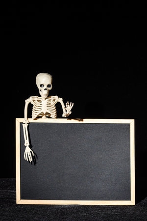 Funny human skeleton holding an empty black blank on a black background, copy space, mockup, vertical frame. Halloween Greeting Cardの写真素材