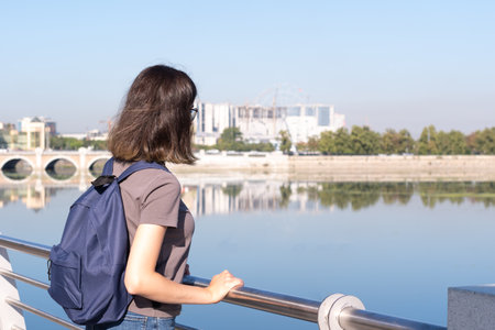 A rear view of a young curly haired college student dressed in a T shirt and jeans and a blue backpack Standing in a park area against the background of the river and the city in the daytimeの写真素材