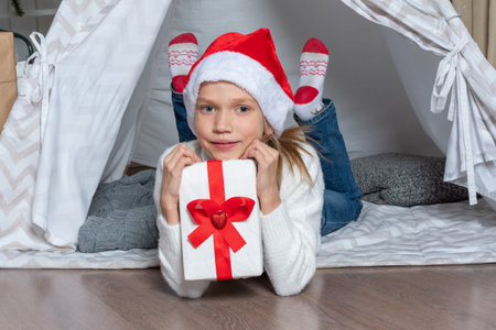 A girl in a Santa hat holds his big gift box for Christmas New Year lying in a childrens tent wigwam in the nursery. Christmas gifts for children. Family Christmas conceptの写真素材