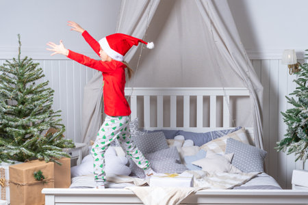 Happy funny baby girl in pajamas and Santa hat jumps in bed on Christmas morning. Children are enjoying the Christmas holiday. The child is having fun.の写真素材