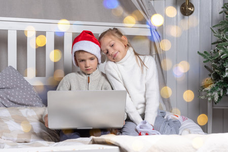 Happy kids in red Santa hats with a laptop on Christmas Eve at home. A boy and a girl online congratulate relatives sitting on the bed. Family x-mas eve time vacation.の写真素材