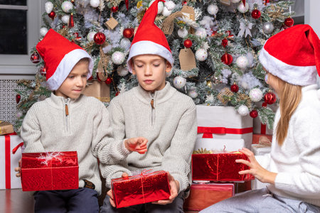 Cute happy excited children, boy and girl in santa hats unwrap christmas present box in beautiful room. Sister and brother give Xmas gifts near decorated tree. Family Christmas and New Year holidayの写真素材