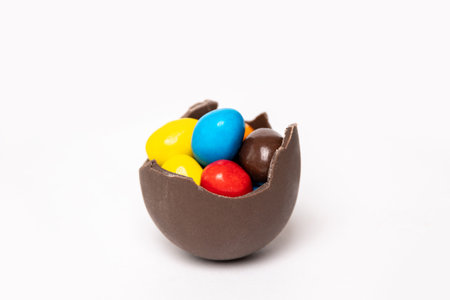 Cracked chocolate easter egg with colorful small round candies on white background, copy space, side view. Chocolate treat for kids. Easter concept.の写真素材