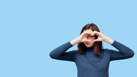 A young beautiful student girl in glasses, in casual clothes, makes a heart shape with her hands instead of eyes, smiles, isolate, blue background, banner. Love, relationships, Valentine's Day.の写真素材