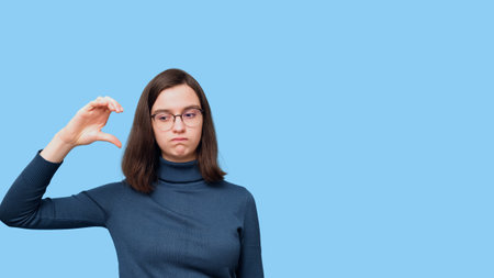 A sad student girl with glasses in a sweater showing a gesture of half a heart on a blue background. A symbol of loneliness. The search for love. To need love. Valentine's Dayの写真素材