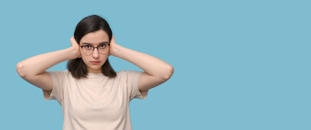 Portrait of a serious student girl in glasses covering her ears with her hands in isolation on a...