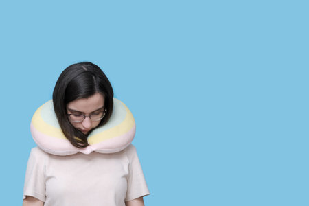 Portrait of a student girl in glasses sleeping in a travel pillow on a blue background, copy space. A woman at rest with a pillow for her neck. The concept of travelの写真素材