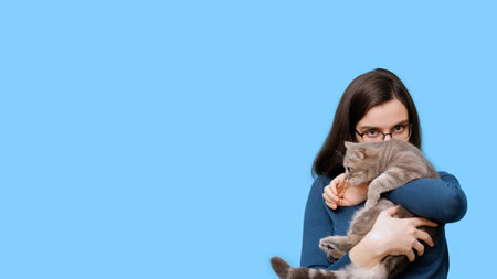 A cheerful student girl hugging a naughty gray striped kitten on a blue background, banner, copy space. A girl strokes a cat. Taking care of pets. gift. love, relationships. Antistressの写真素材
