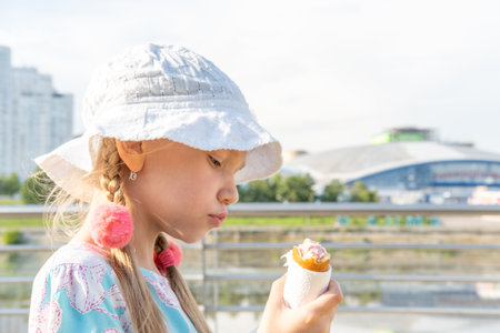 Hungry kid eating street food on the beach in summer. A girl in a hat eating fried sausages. Happy kids eating fast food. Fast high-calorie street junk food.の写真素材
