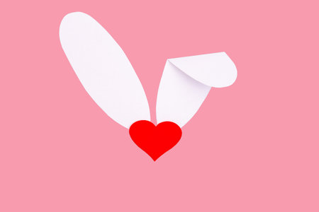 creative greeting card. Imitation of the Easter bunny. A red heart instead of a nose and white paper ears on a pink background. The minimum concept of a spring holiday.の写真素材