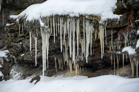 Drops melting water from roof fall onto snow and frozen icicles next to stone in spring. Icicles and...