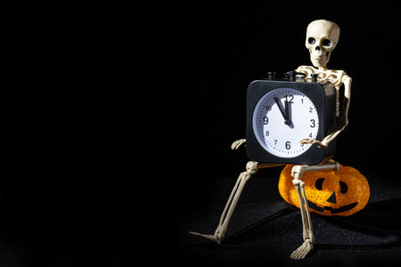 A skeleton sitting on a letter-shaped lamp with a grin and holding an alarm clock on a black background. Time of the Dead.の写真素材