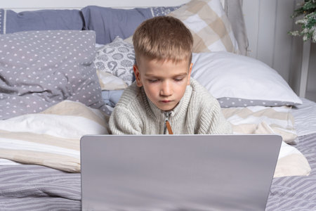 the child is a boy lying on the bed and looking at a laptop. Elementary school student doing...