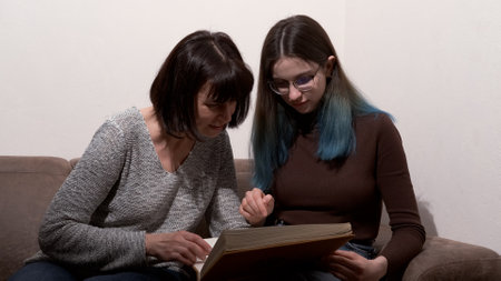 Mother and daughter watch family photos, remember pleasant moments. Beautiful woman and her teenage daughter are looking through photos and smiling while sitting on couch at home.の写真素材