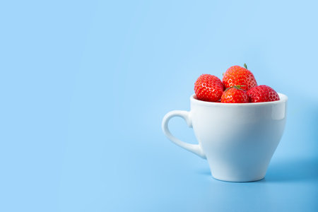 A glass white cup full of red ripe strawberries isolated on blue background, copy of space. Summer healthy drink. Summer berry tea. Healthy fruit drinks. Berry background. Harvesting strawberriesの写真素材