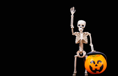 A human skeleton greeting, waving, holding a Halloween orange pumpkin on a black background. Halloween Greeting Cardの写真素材