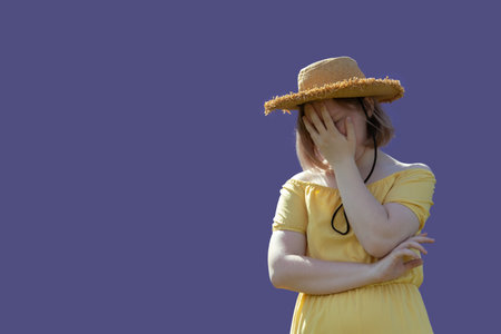 A frustrated teen girl in a hat and dress covers her face with her hands on a purple background. Mental health concept.の写真素材