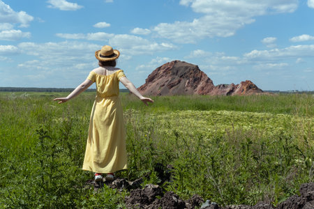 Girl in a straw hat and yellow dress among the fluffy grass in a field, rock, rear view. Summer time. Village vacation. Time to travelの写真素材