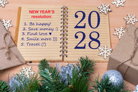 2028 New Year's Resolution - Hand holding a pen and writing down plans for the new year: be happy, find love, save money, smile, travel on the table with gifts, snowflakes and fir branches with balls.の写真素材