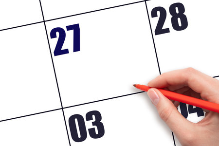 Calendar date 27. A hand holding a red pen is about to write text on an empty white calendar page with the date 27 without a month. Planning or scheduling concept.の写真素材