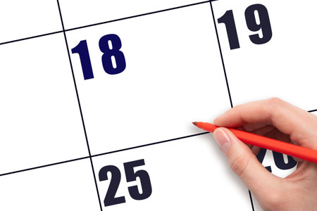 Calendar date 18. A hand holding a red pen is about to write text on an empty white calendar page with the date 18 without a month. Planning or scheduling concept.の写真素材