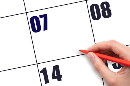 Calendar date 7. A hand holding a red pen is about to write text on an empty white calendar page with the date 7 without a month. Mockup. Save the date. Holiday. Planning or scheduling concept.の写真素材