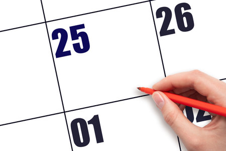 Calendar date 25. A hand holding a red pen is about to write text on an empty white calendar page with the date 25 without a month. Planning or scheduling concept.の写真素材