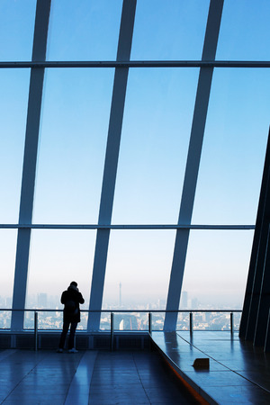 Young man standing by the window in the Sky Garden, the highest public garden in London, taking pictures of the city skyline on mobile phone.のeditorial素材