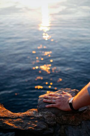Close up shot of men's left hand on a rock, wearing a simple wedding band and a watch. Sea, sunset and light trails on the background.の写真素材