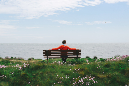 Young man in red jacket sitting on a bench, looking at the seaの写真素材