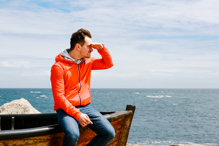 Young man in jeans and red jacket sits on the edge of a boat by the sea, looking at the distance, hand on foreheadの写真素材