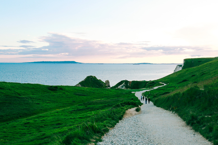 People walking on a path amongst greenery towards the famous Durdle Door on Jurassic Coast, Dorset, UK at dusk.の写真素材