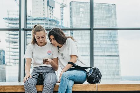 Two young women sitting by the window Sky Garden, London, looking up information on mobile phone. Sky Garden, the highest public garden in London.のeditorial素材
