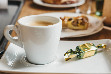 Closeup of a fresh cup of coffee on a saucer, chocolate in foil next to it, plate with cake on the background, selective focus.の写真素材