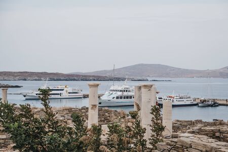 Delos, Greece - September 20, 2019: Ruins on the Island of Delos, archaeological site near Mykonos in the Aegean Sea Cyclades archipelago, tour boats docked on the background.のeditorial素材