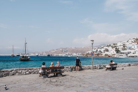 Mykonos Town, Greece - September 23, 2019: People relaxing on a bench by the water on a sunny day in Hora, also known as Mykonos Town, the capital of popular Greek island Mykonos.のeditorial素材
