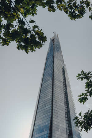 London, UK - August 25, 2020: Low angle view of top of The Shard against the sky, through the trees....