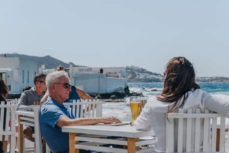 Mykonos Town, Greece - September 24, 2019: Middle aged couple relaxing at the outdoor tables of a restaurant by the water in Hora (also known as Mykonos Town), capital of the Mykonos island.のeditorial素材