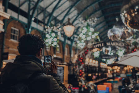 London, UK - November 23, 2021: Man taking photos of Christmas decorations in Covent Garden Market, one of the most popular tourist sites in London, on the mobile phone, focus on the screen.のeditorial素材