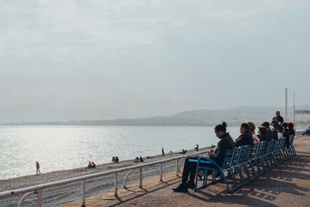 Nice, France - March 10, 2022: People relaxing on blue chairs on The Promenade des Anglais in Nice, a famous tourist destination on the French Riviera, on a sunny spring day.のeditorial素材