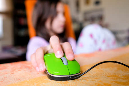 In the foreground is a childs hand on a computer mouse. In the background, a girl is sitting on a chair. Blurred background, colorful photoの写真素材
