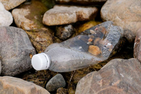 A transparent plastic bottle with a white cap lies on the river bank among the stones