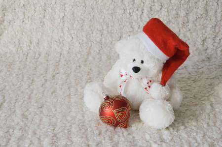 A white toy bear in a red Christmas hat and a beautiful fir ball for decoration. Background for winter holidays, Christmas and New Year.の写真素材