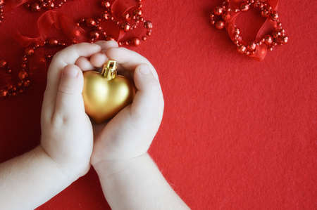 a child holds a toy golden heart on a red background. red beads for decorating a festive fir tree. decoration for Christmas and New Year.の写真素材