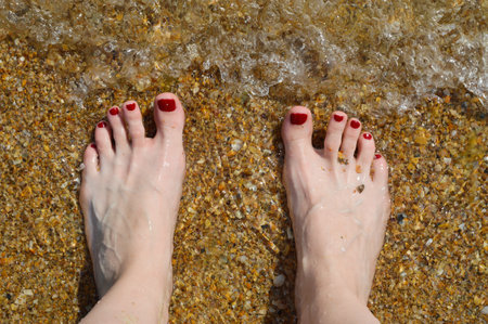 women's feet are standing on the seashore. sea, sand, summer. clear water. nails painted with red varnish. background for the design.の写真素材